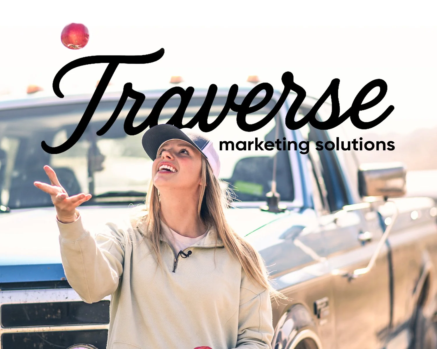 Young woman in casual clothing and baseball cap gesturing with her hand, standing in front of a truck, with text overlay reading 'Traverse marketing solutions'.