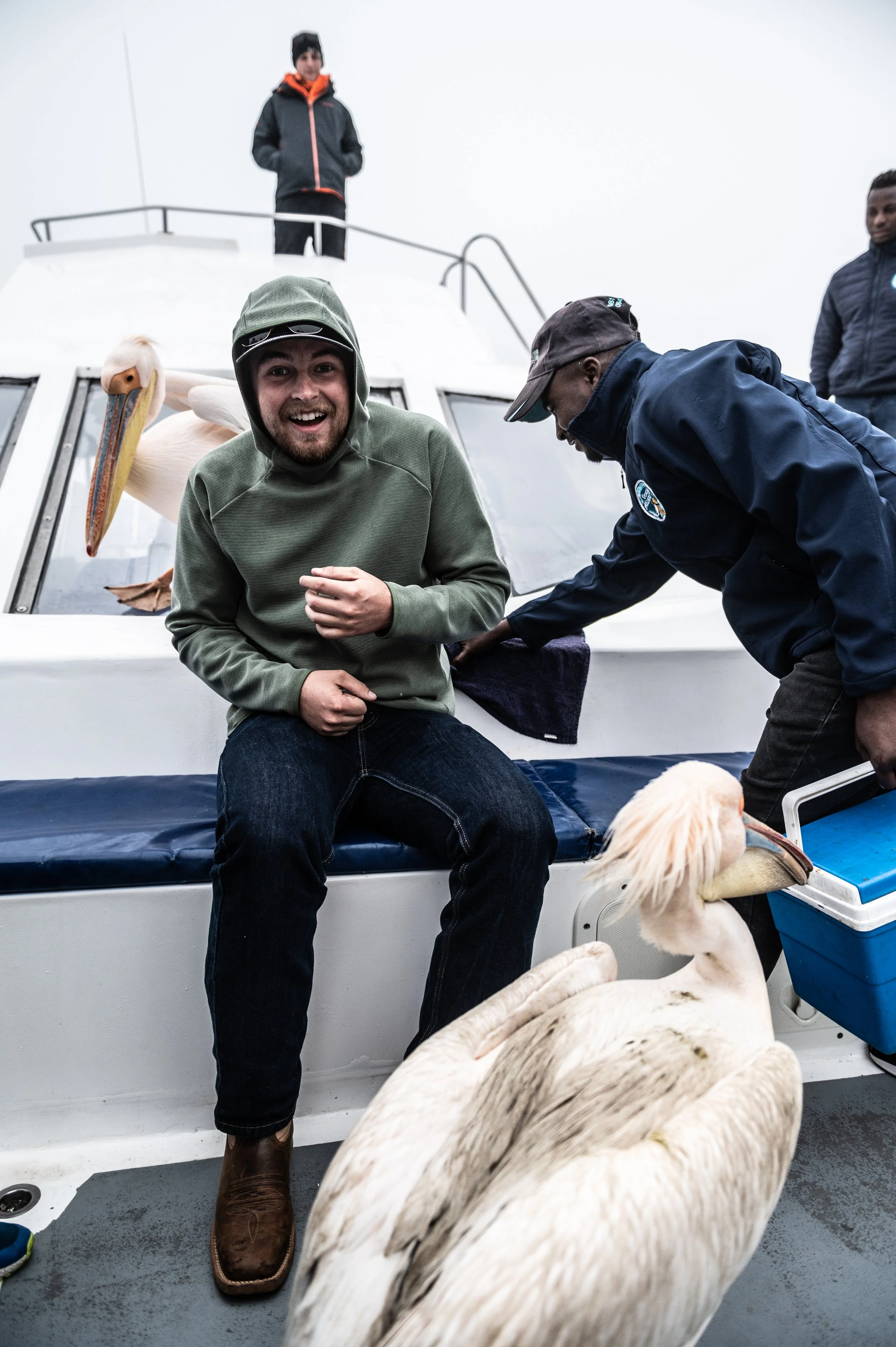Group of people on a boat with swans, one person smiling at the camera, others engaged in different activities, overcast sky.