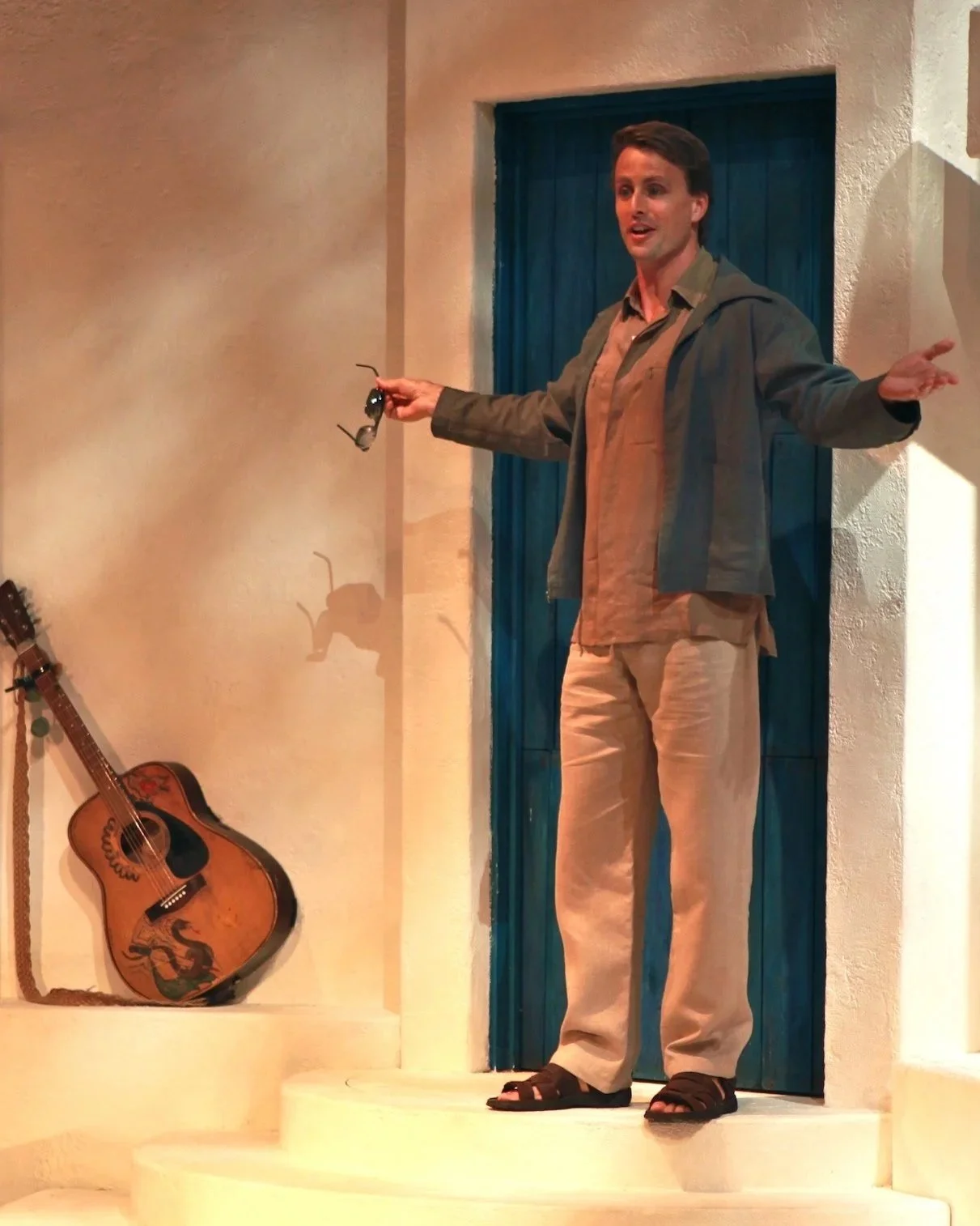 A man standing on a small white staircase, holding sunglasses in his right hand, in front of a blue door with a beige wall, with a guitar leaning against the wall on his left.