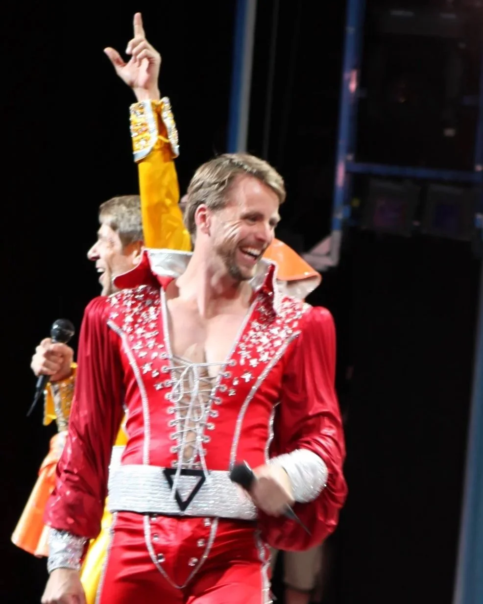 Performer in red costume dancing with a microphone, surrounded by people in colorful costumes holding microphones, on stage with curtains in the background.