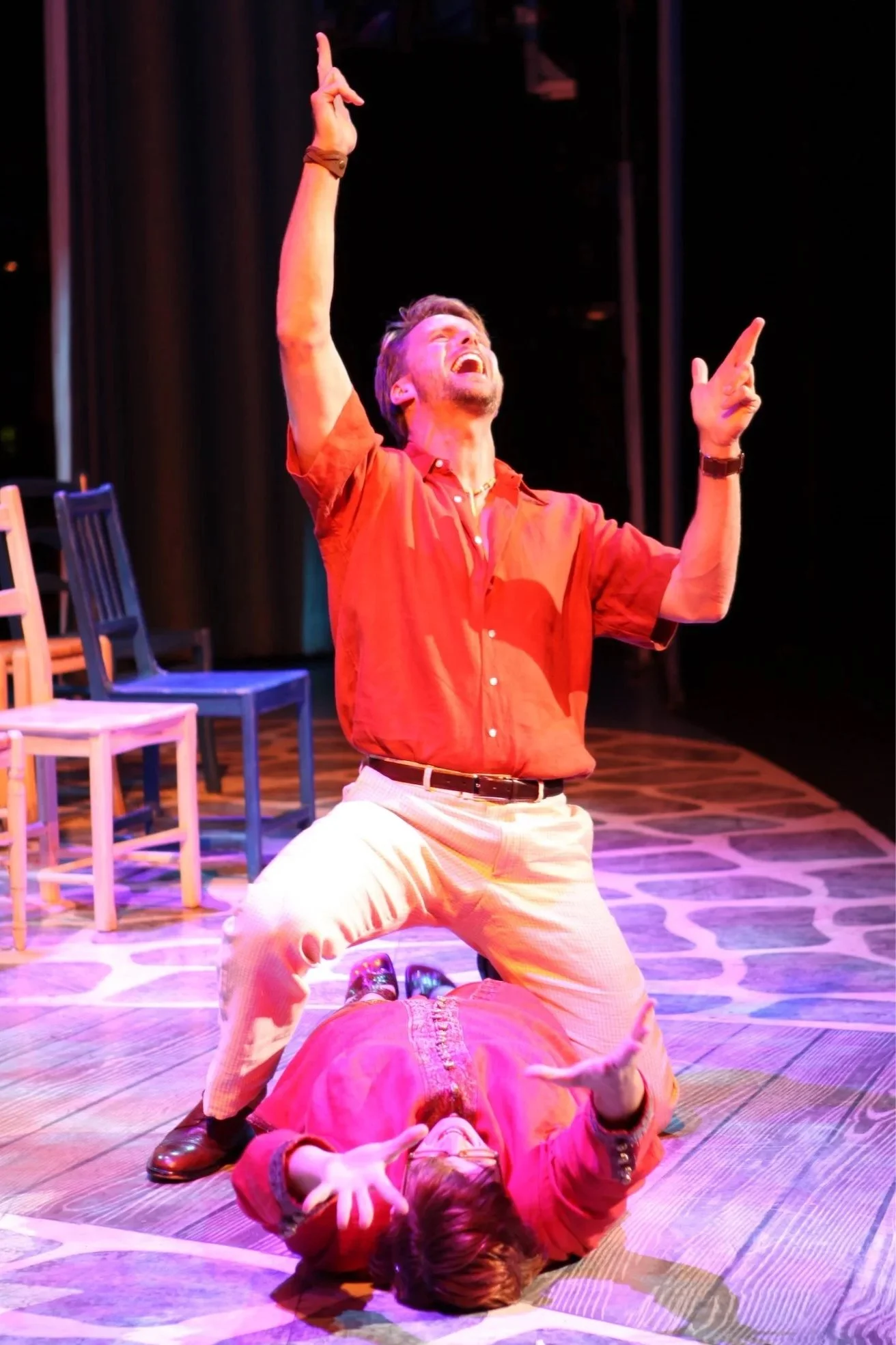 A theatrical scene with a man kneeling over a woman on the stage, both in mid-dramatic expression. Several chairs are arranged on the stage in the background, with stage lighting casting colorful hues.