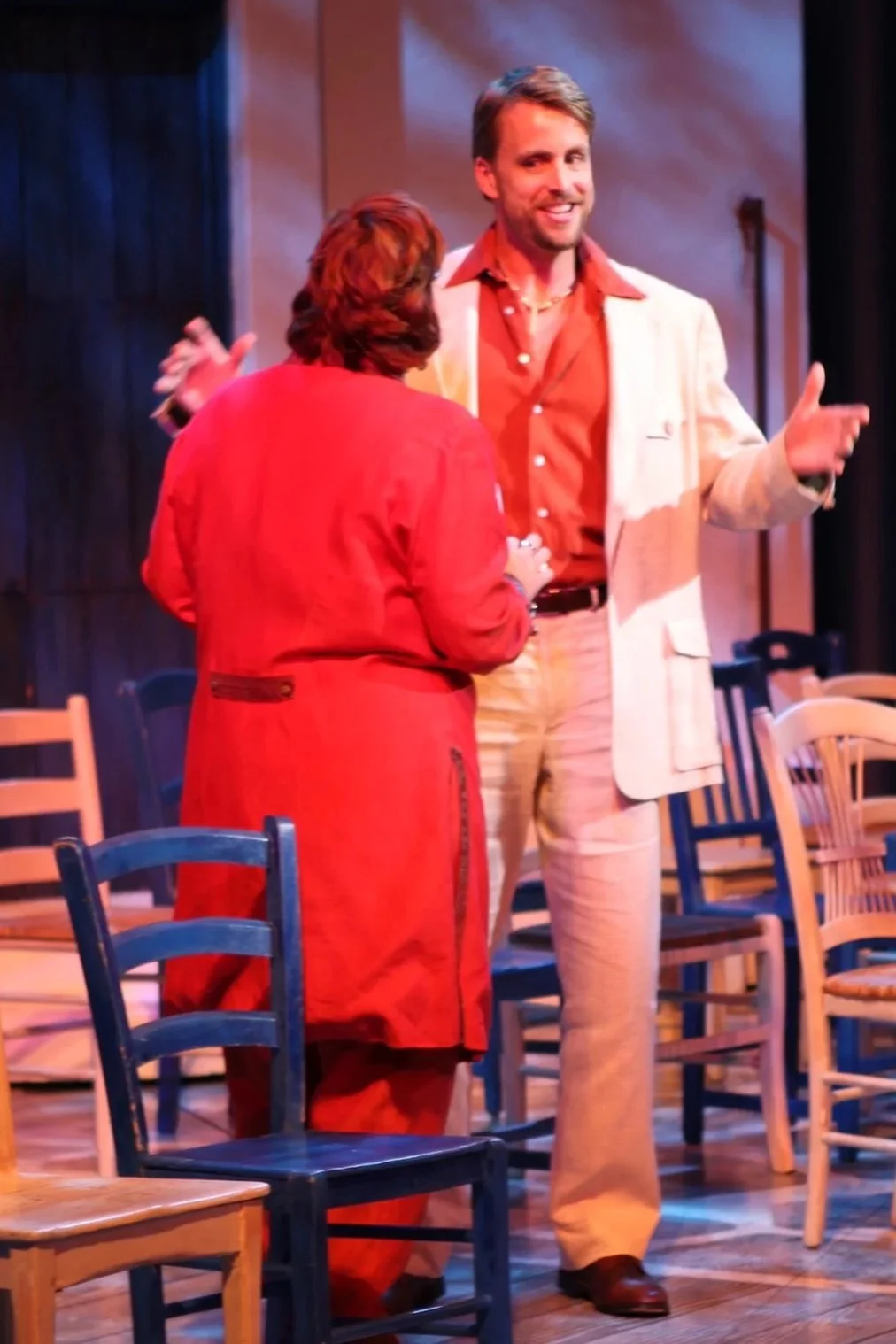 Two actors on stage during a theatrical performance. The woman, wearing a red dress, and the man, dressed in a cream-colored suit, are engaged in a conversation amid scattered chairs. The stage has a simple backdrop with beige walls and a wooden floo