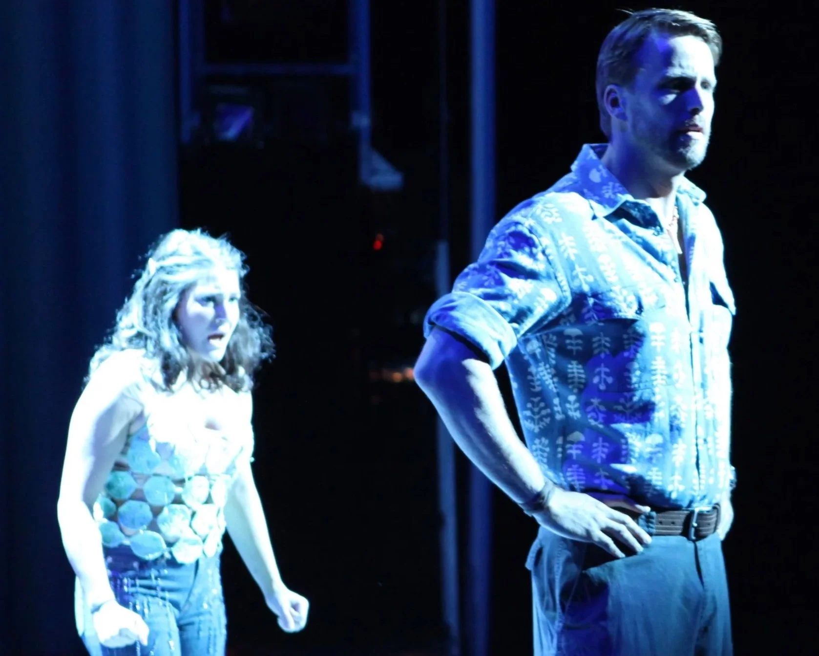 Two actors on stage, one man and one woman, under blue lighting. The woman has curly hair and worn jeans, the man has short hair, a beard, and is wearing a patterned shirt with rolled-up sleeves.