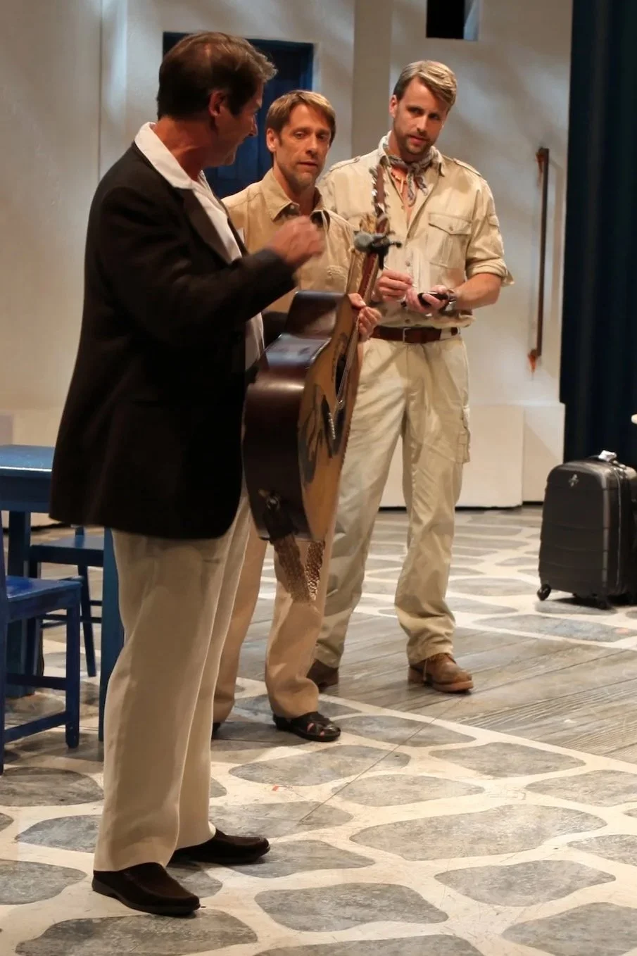 Three men on stage, one holding a guitar, in a theatrical setting with blue chairs, small tables, luggage, and a white backdrop, preparing for a scene.