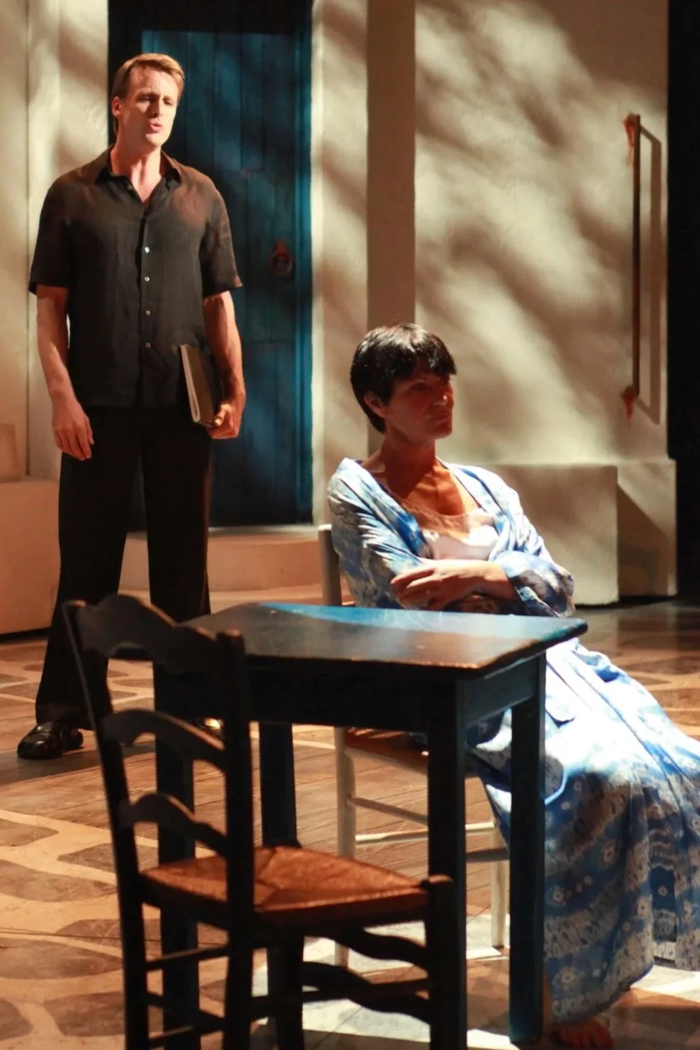 Two people on a stage set that resembles a Mediterranean-style courtyard, with a woman sitting at a table and a man standing nearby, holding a notepad.