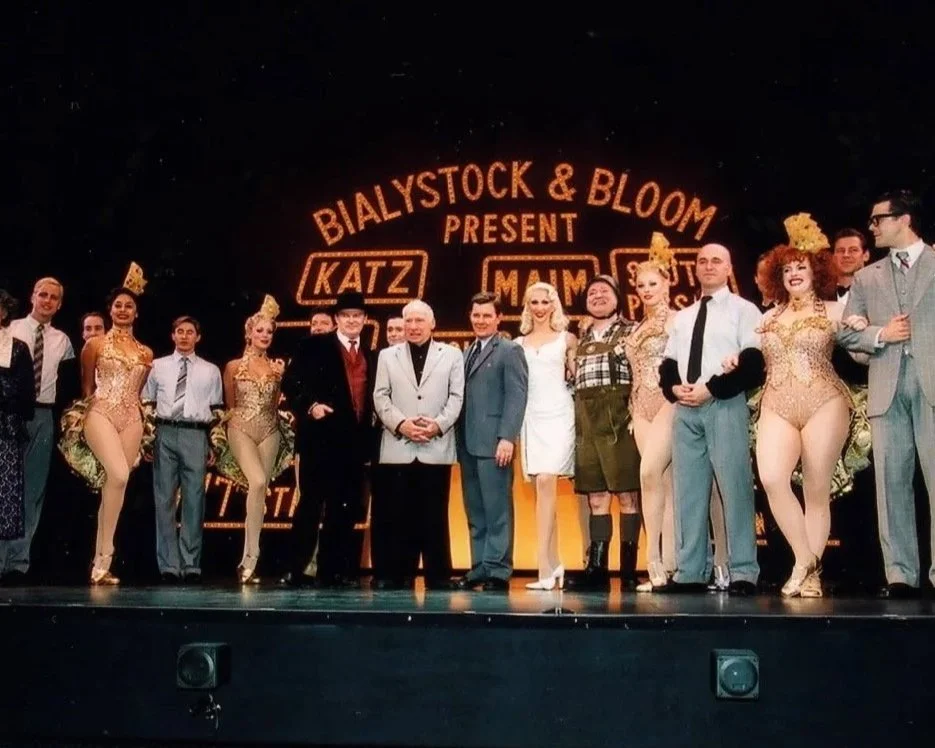 Cast of a musical theater production on stage, dressed in vintage costumes, with a backdrop resembling a city billboard.