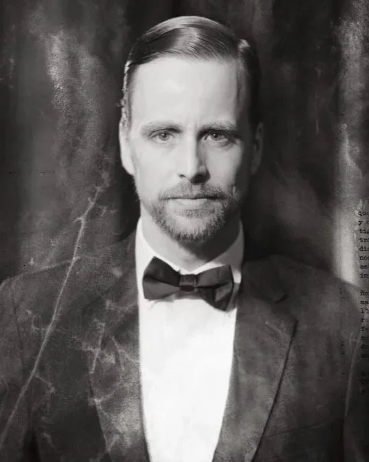 Black and white photo of a man with neatly combed hair and a beard, wearing a tuxedo with a bow tie, against a dark backdrop.