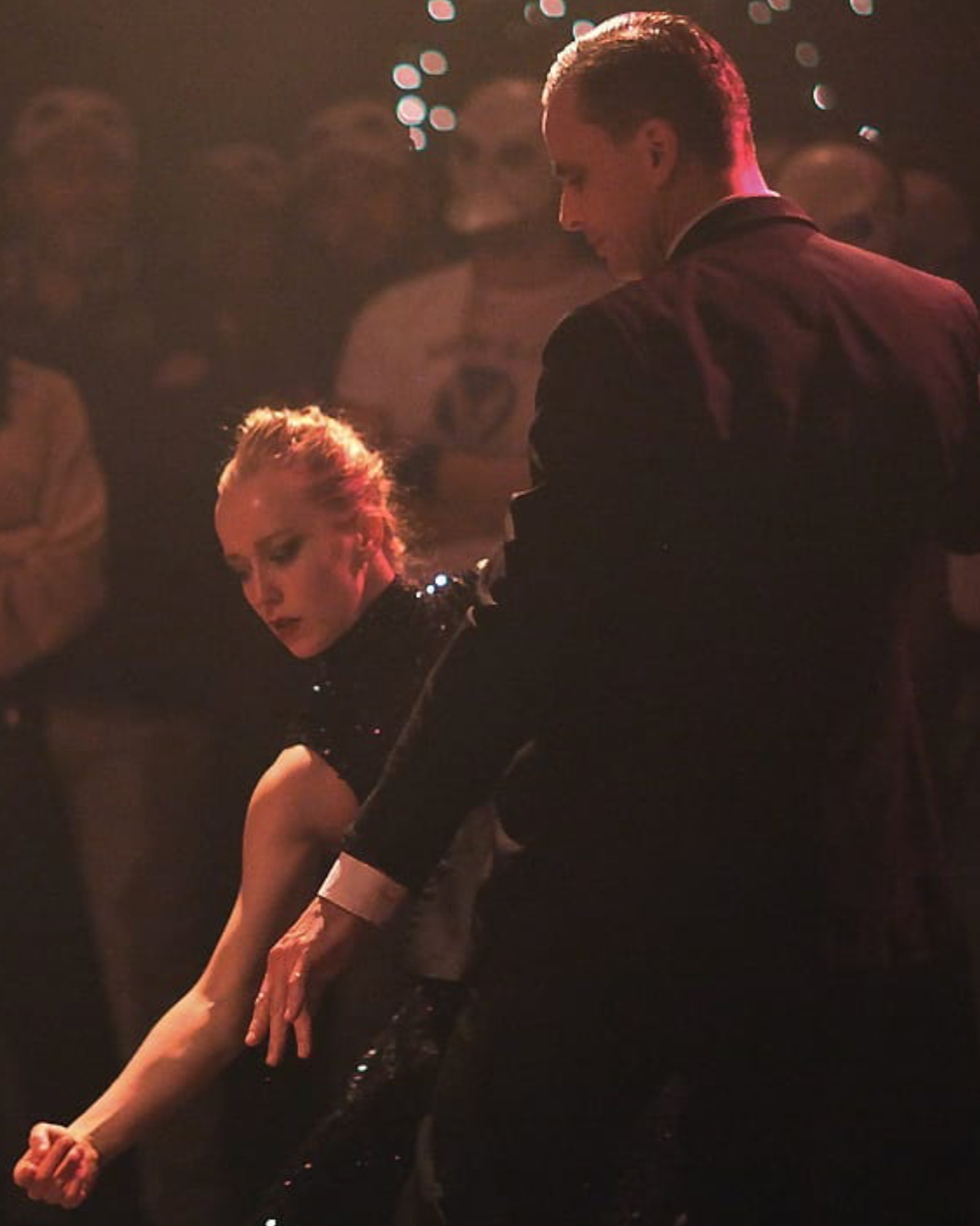 A man and woman dance closely in a dimly lit setting, with the woman wearing a black sequined dress and the man in a black suit, amidst an audience observing intently.