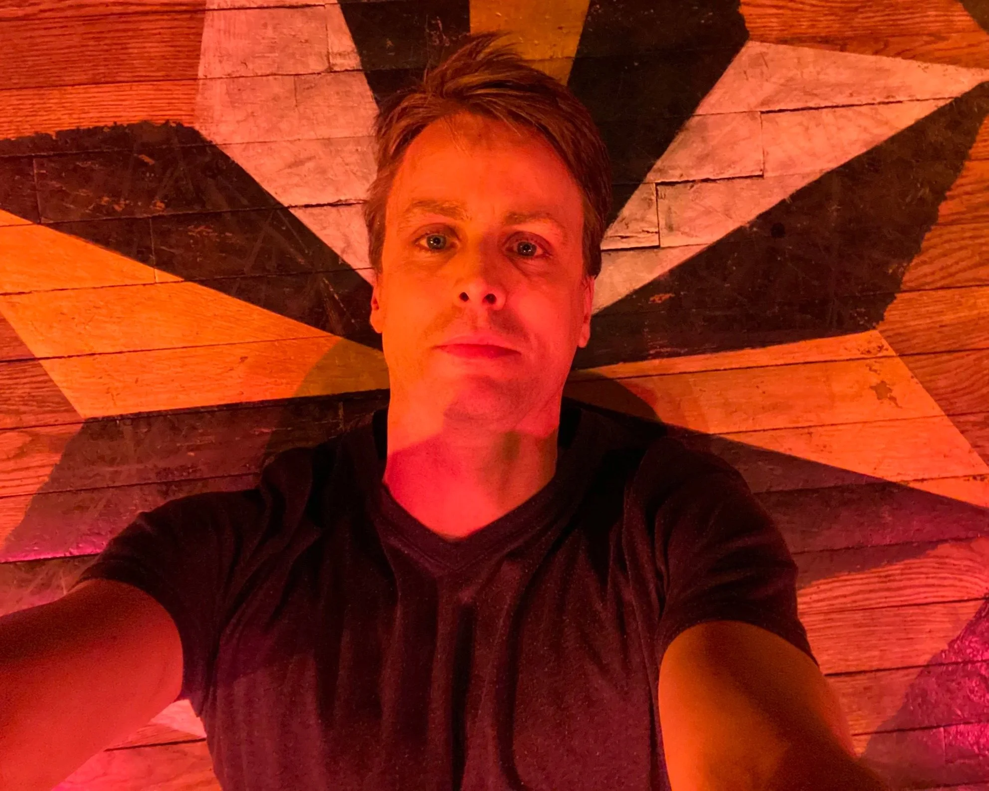 A person taking a selfie on a wooden floor with a geometric star pattern in pink, black, and beige colors, illuminated by warm-colored lighting.