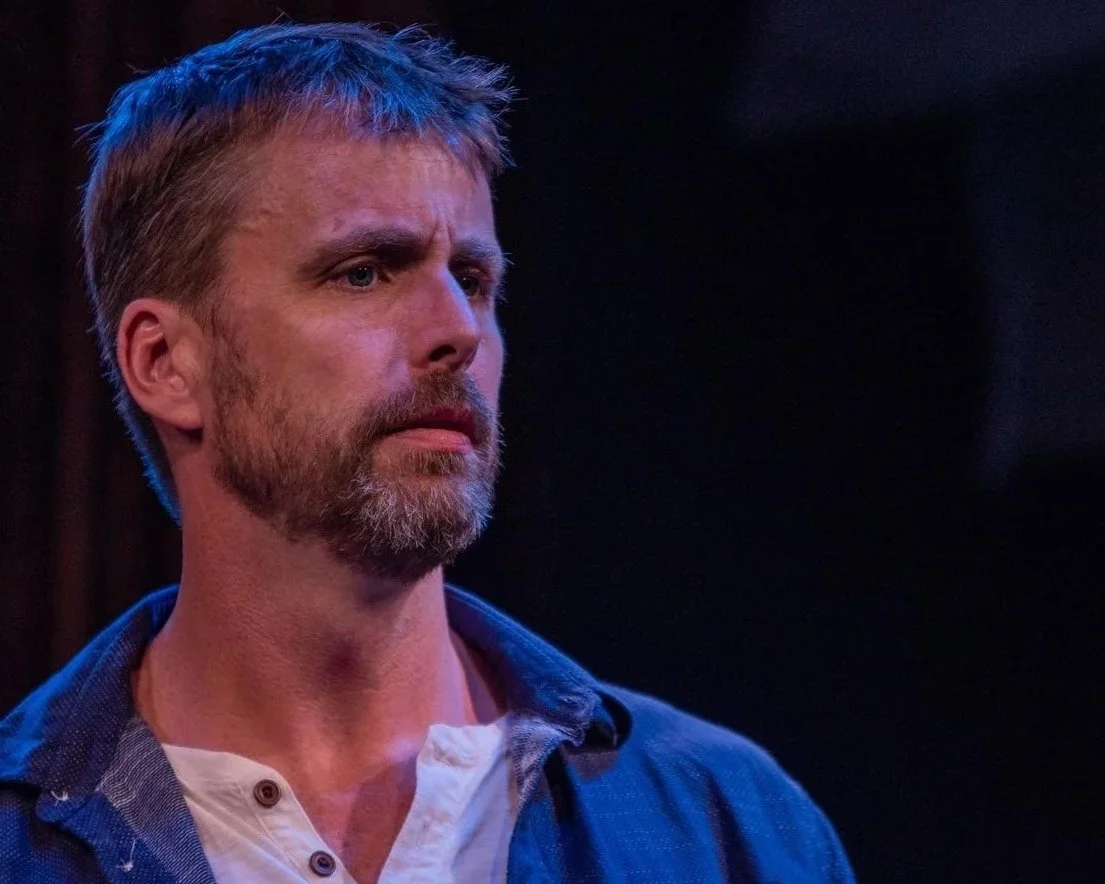 A man with a beard and mustache, wearing a blue shirt over a white undershirt, with a serious expression, standing in a dimly lit environment.