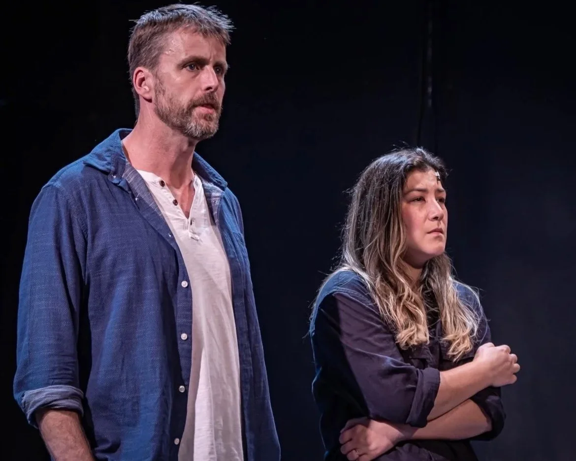 A man and a woman standing on a dark stage, appearing serious or contemplative.