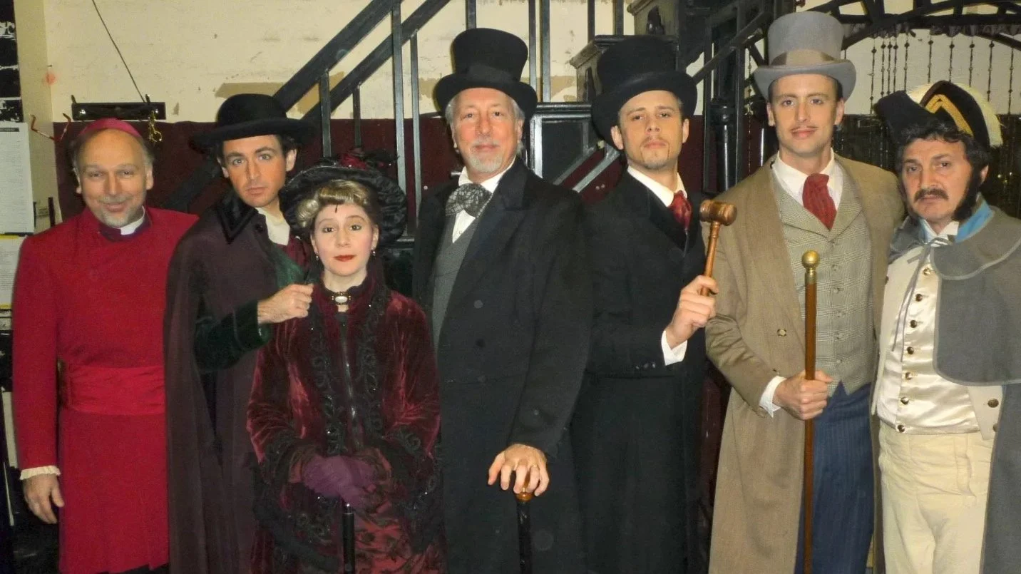 Group of eight people dressed in Victorian-era costumes, posing in front of a staircase inside a building.