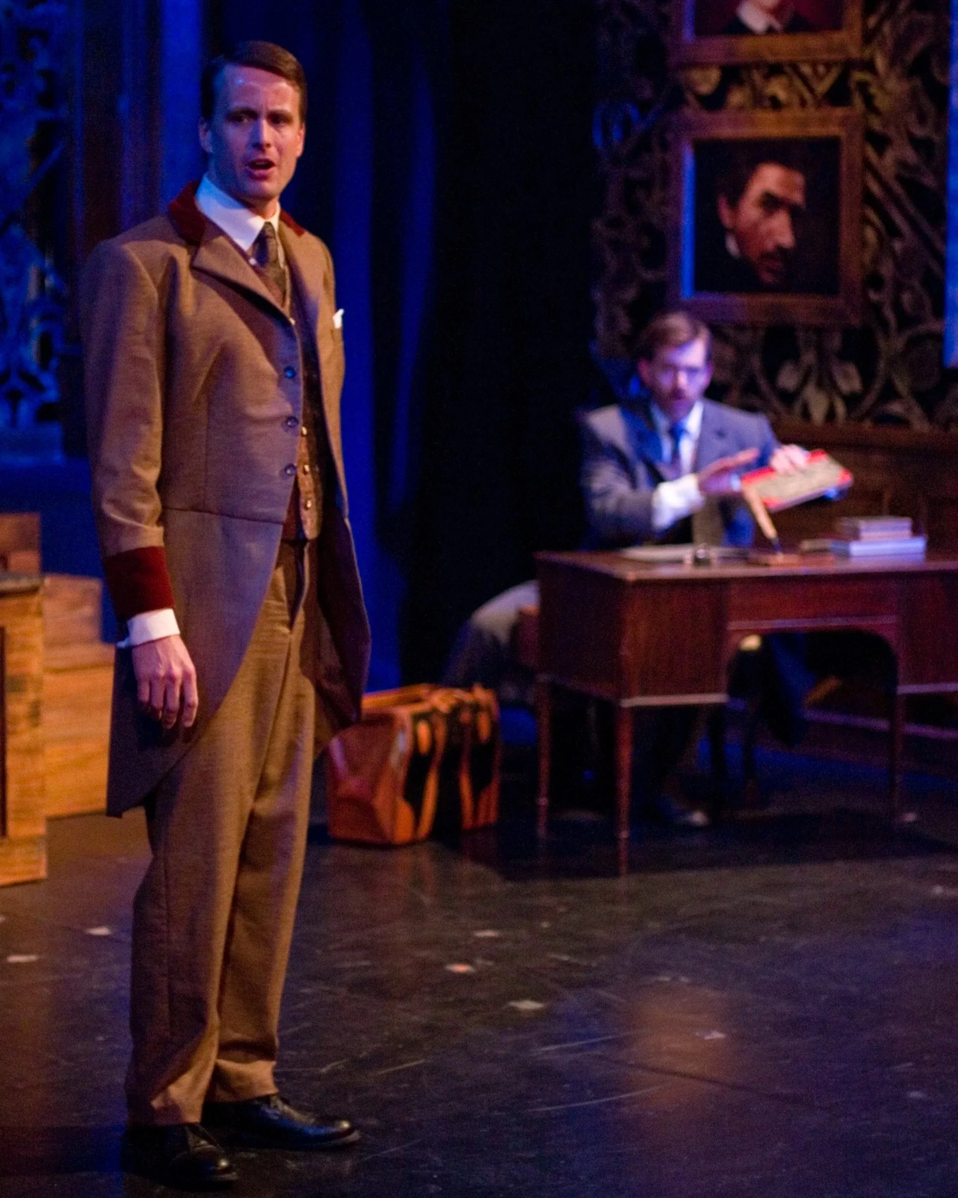 A man in vintage-style suit standing on a stage with a dark ornate background, with a person sitting at a desk in the background holding an object. Portraits are hanging on the wall behind.