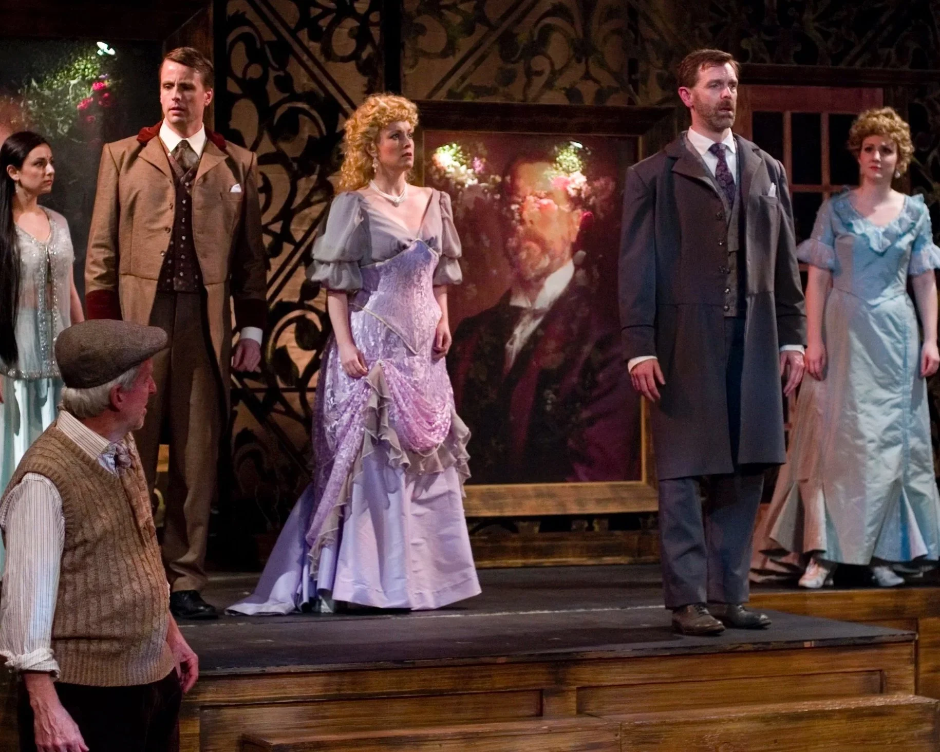 The cast of a theatrical performance on stage, dressed in period costumes, with a large framed portrait in the background.