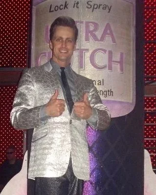 A man in a shiny silver suit giving a thumbs up, standing next to a giant spray can labeled "Ultra Catch" with a sign above that says "Lock it Spray." The background features a red patterned backdrop.
