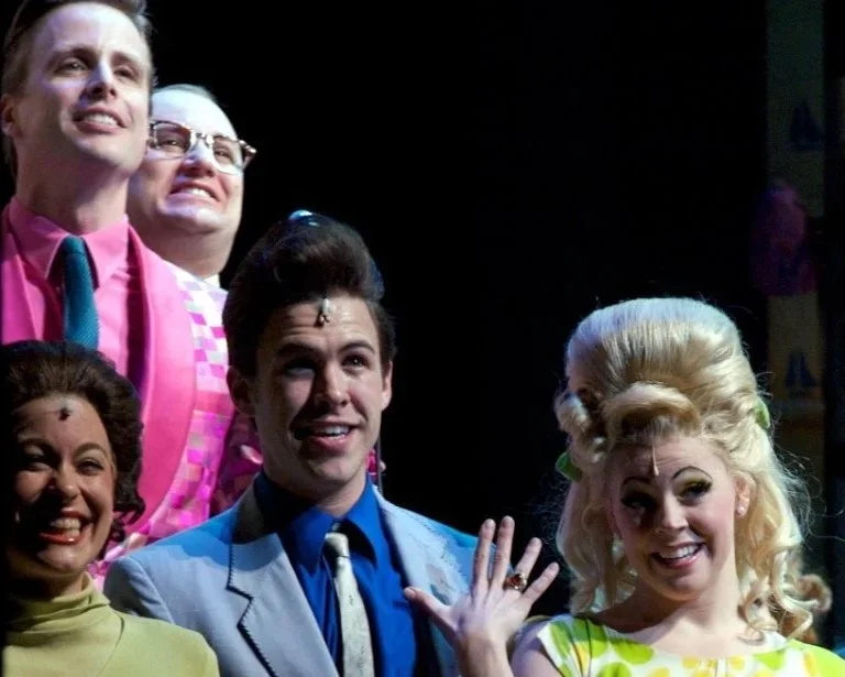 Group of people in colorful vintage outfits posing and smiling, likely from a theatrical or entertainment setting.