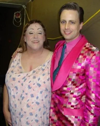A woman and a man standing close together, smiling, in a backstage or dressing room area with a dark wall and yellow cables in the background.