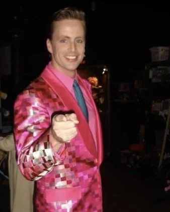 A man in a shiny pink checkered suit pointing at the camera, smiling, in a dark room or backstage setting.