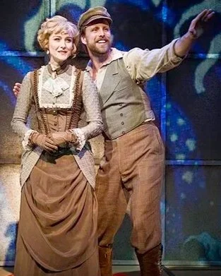 Two actors on stage, dressed in vintage clothing, with the man pointing and smiling, and the woman standing beside him with her hands clasped.