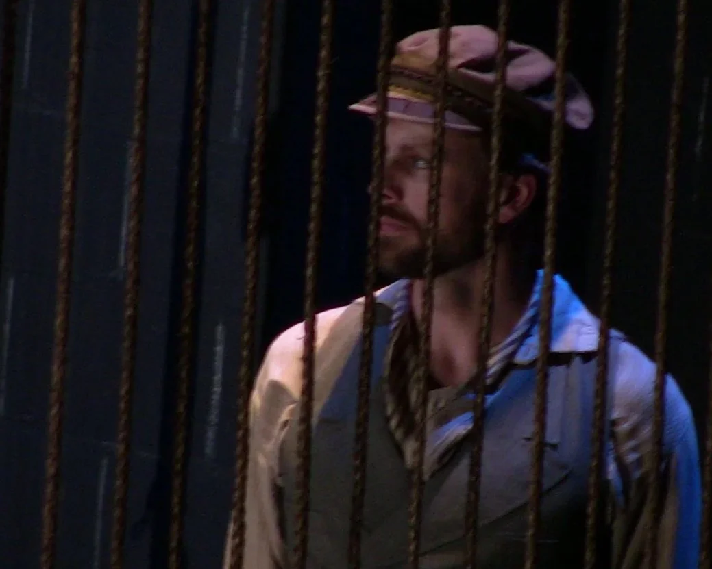 A man wearing a pink hat and striped shirt behind black metal bars, facing slightly to the side in a dark environment.