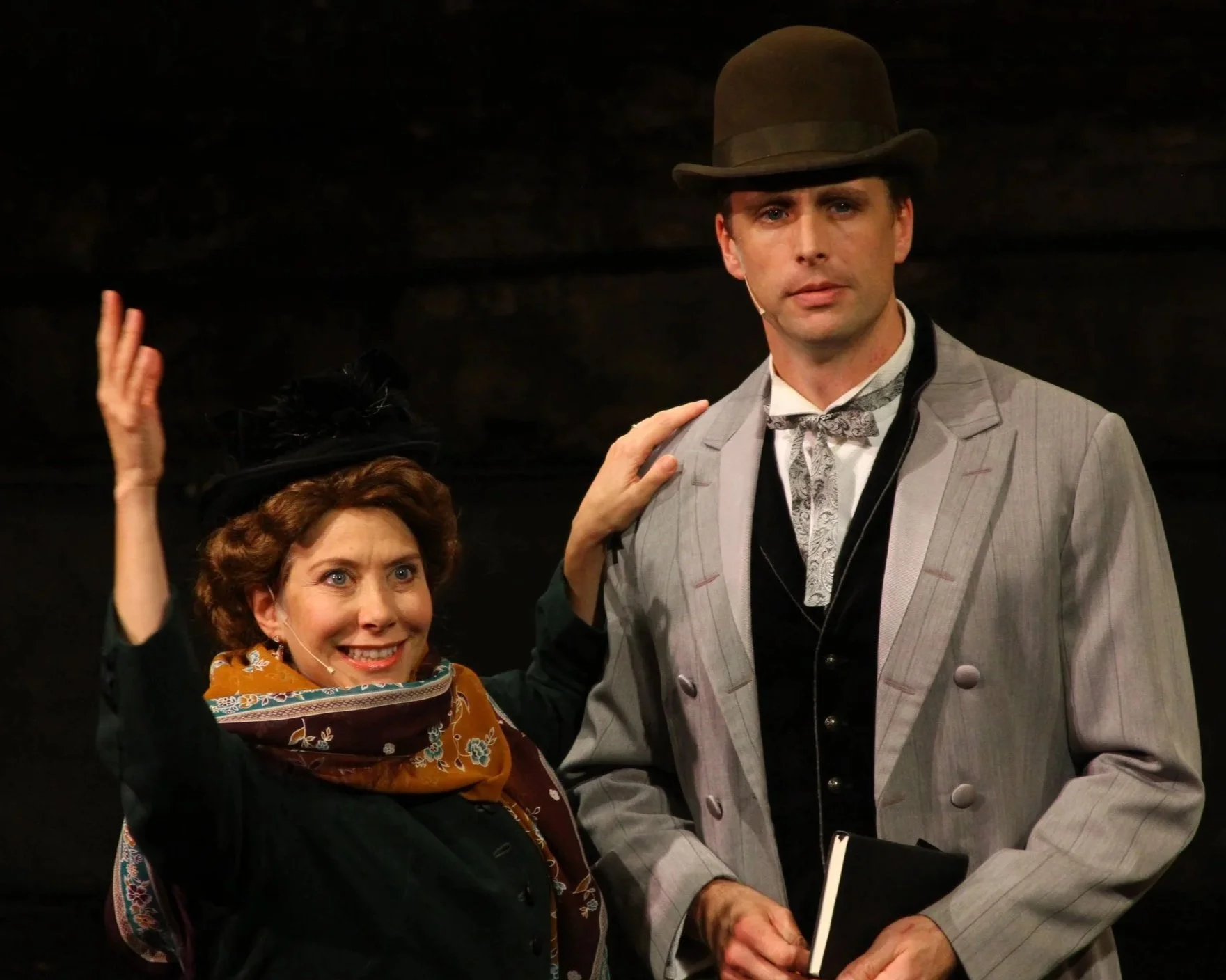 Two actors on stage, one woman with a scarf and a hat talking animatedly and the man next to her wearing a bowler hat and vintage suit, holding a notebook, with another person partially visible on the right.