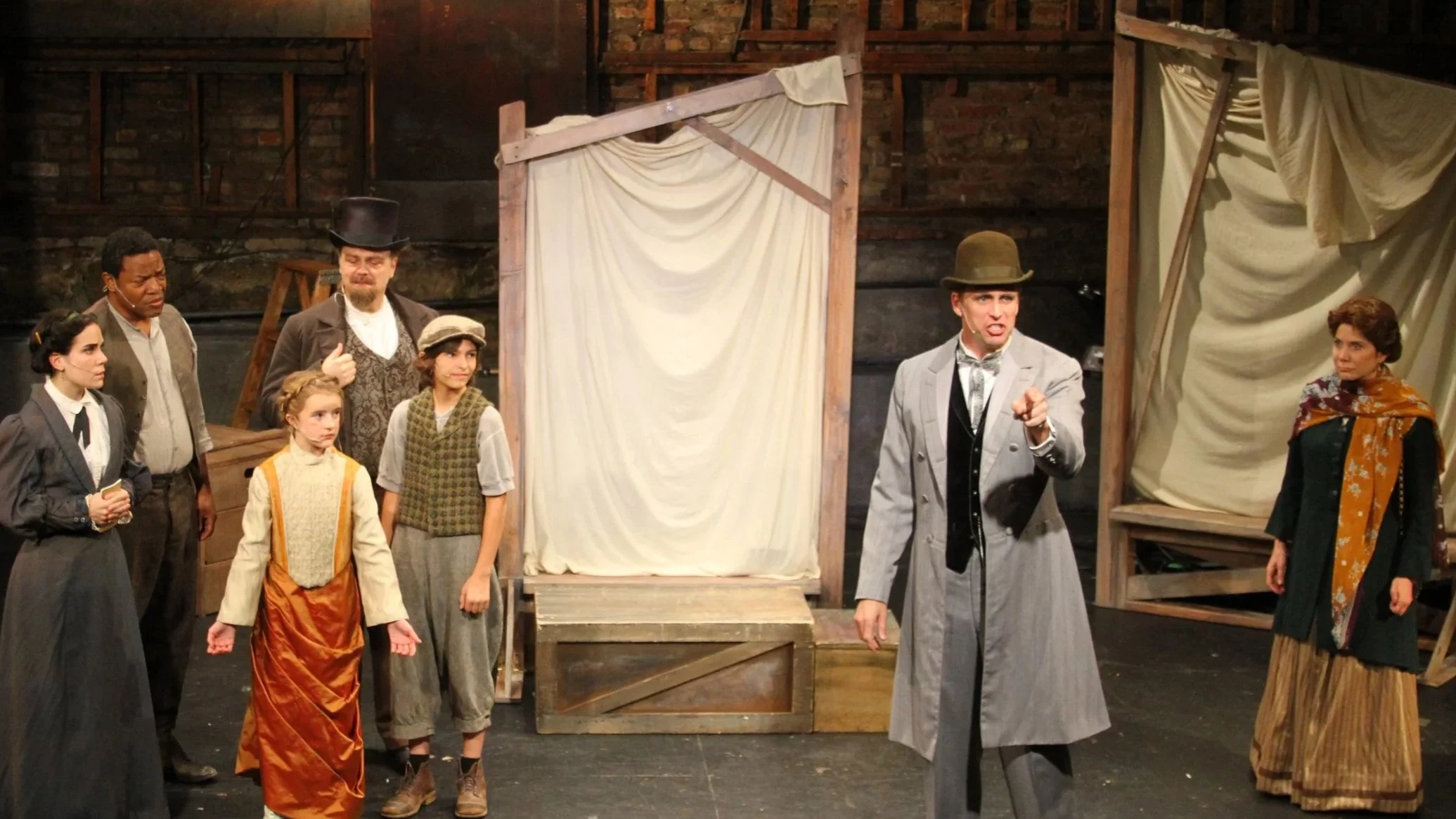Theater scene with multiple actors in period costumes performing on stage with wooden set pieces and curtains.