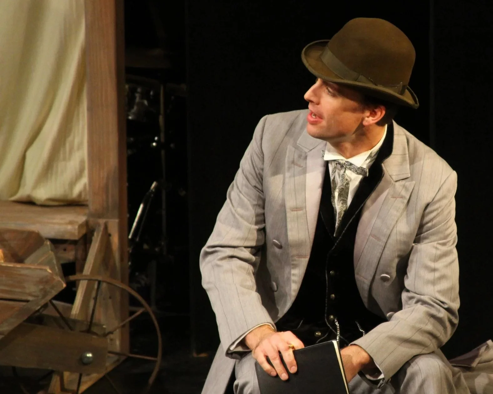 Actor in vintage suit and hat sitting on a wooden box on stage, holding a book, with props like a wheel and wooden structure nearby.