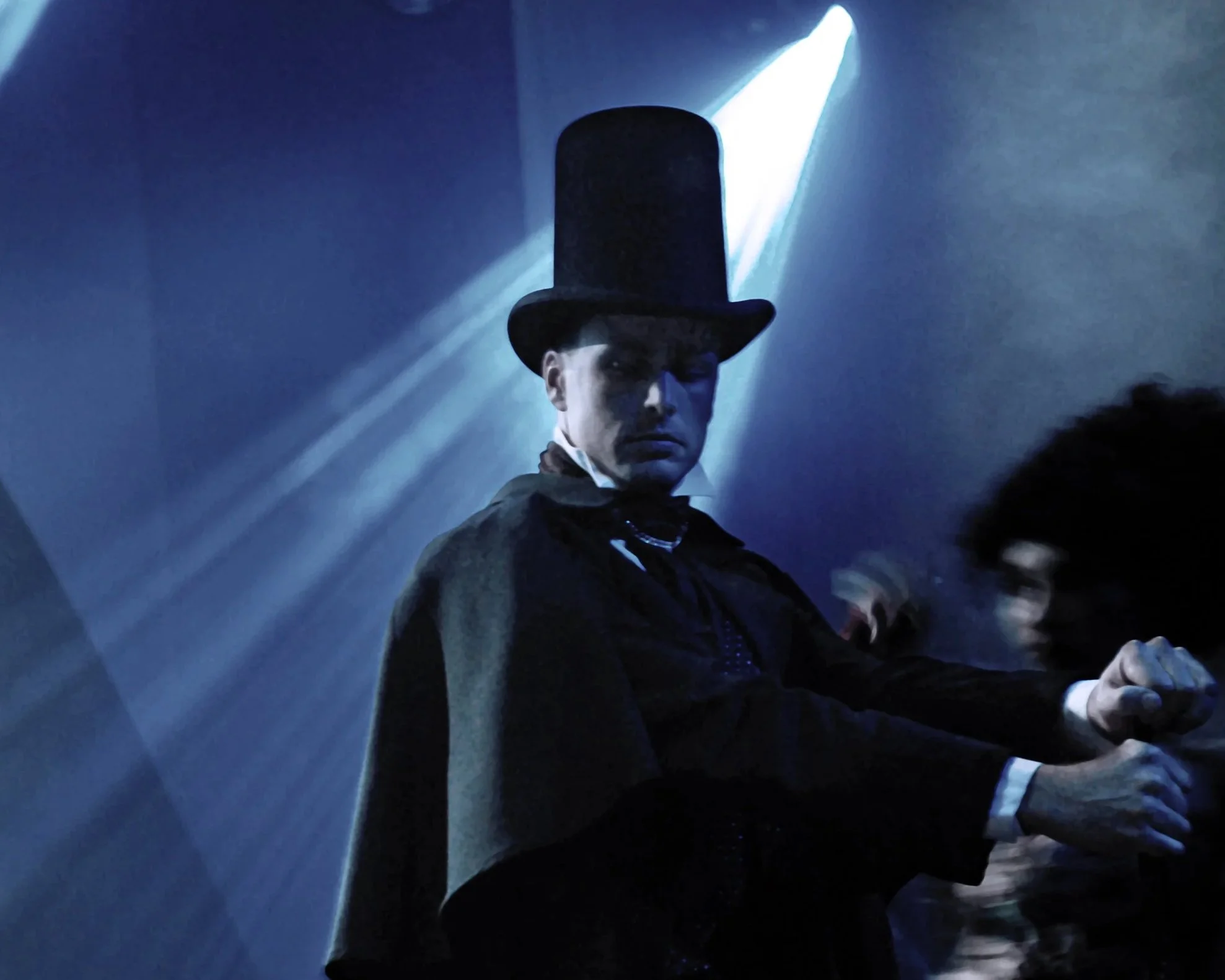 Person dressed as Abraham Lincoln with a tall top hat and cloak, standing under blue stage lighting at a costume event or theater.