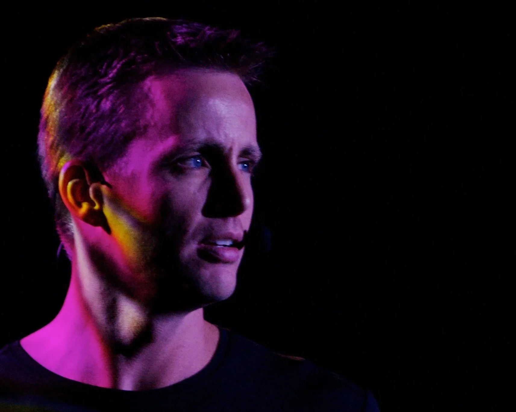 A man with short hair and blue eyes looking to the left, with pink and yellow lighting on his face, dark background.