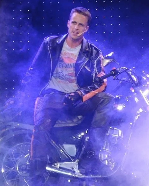 A person sitting on a motorcycle on a stage with blue and white smoke effects, wearing a leather jacket and a graphic T-shirt, with a dark background of small illuminated dots.