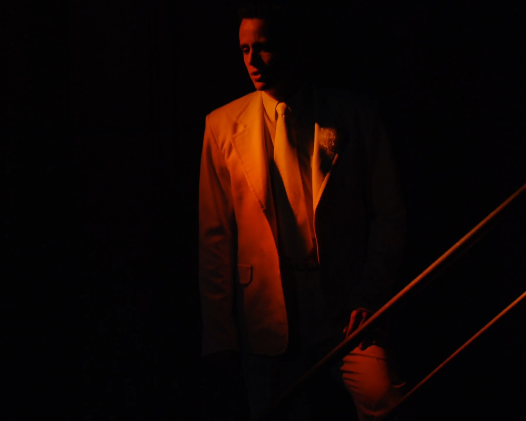 A man in a beige suit and tie stands in a dark room with minimal lighting, creating a dramatic shadowed effect.