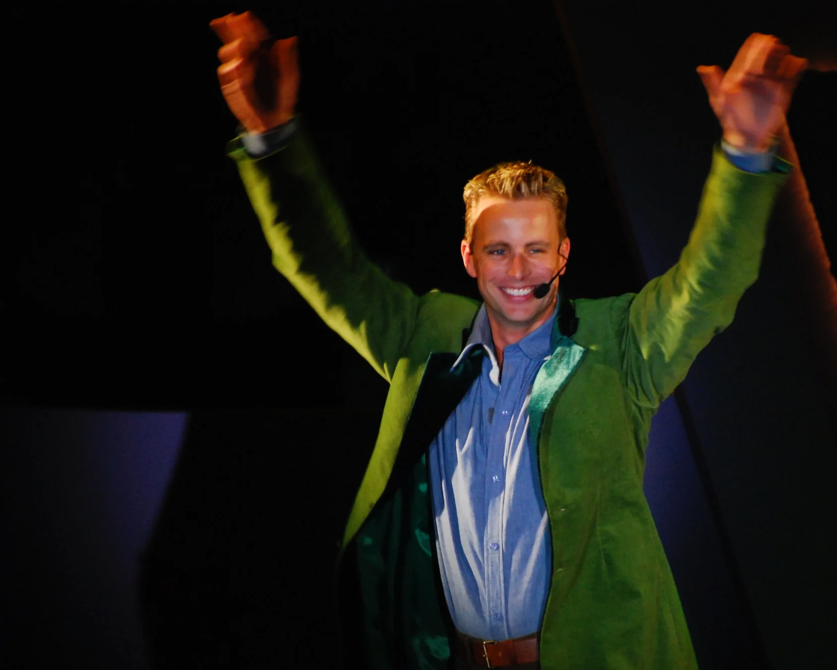 A man in a green jacket and blue shirt smiling and raising both arms, standing on stage with a microphone headset.