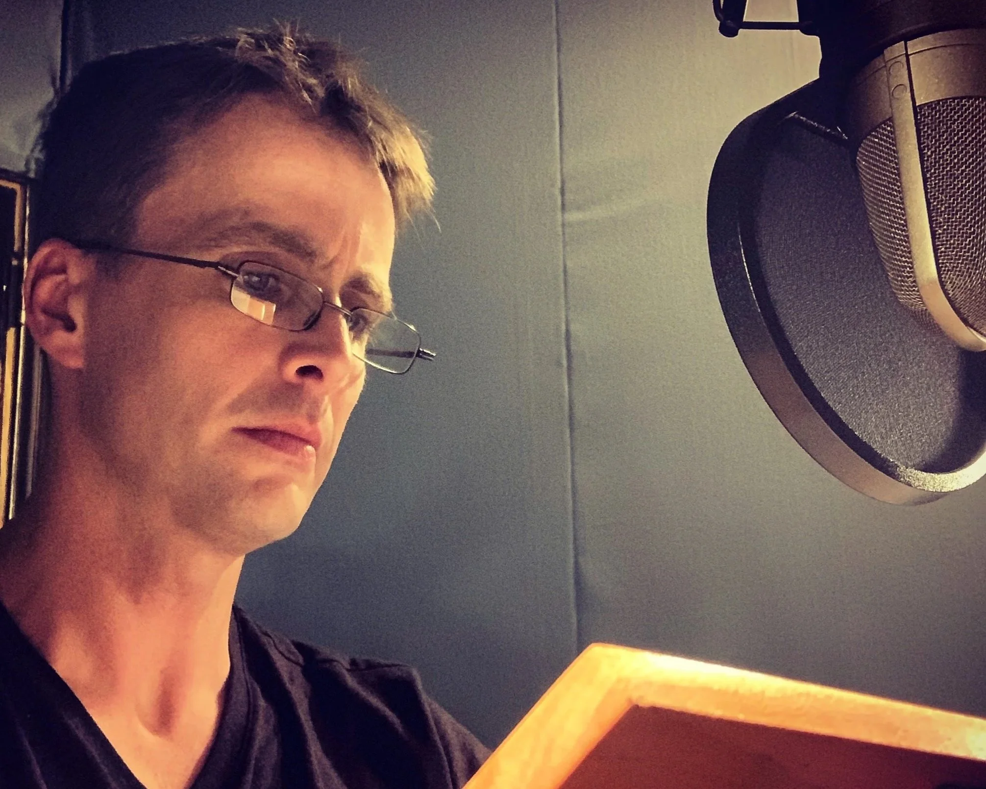 A man wearing glasses is recording vocals in a music studio, standing near a microphone and reading from a sheet of paper.