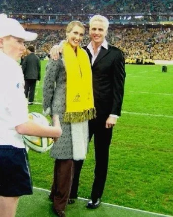 Two women and one man standing on a stadium field, smiling, with a crowd in the background, next to a young girl in a sports uniform holding a rugby ball.