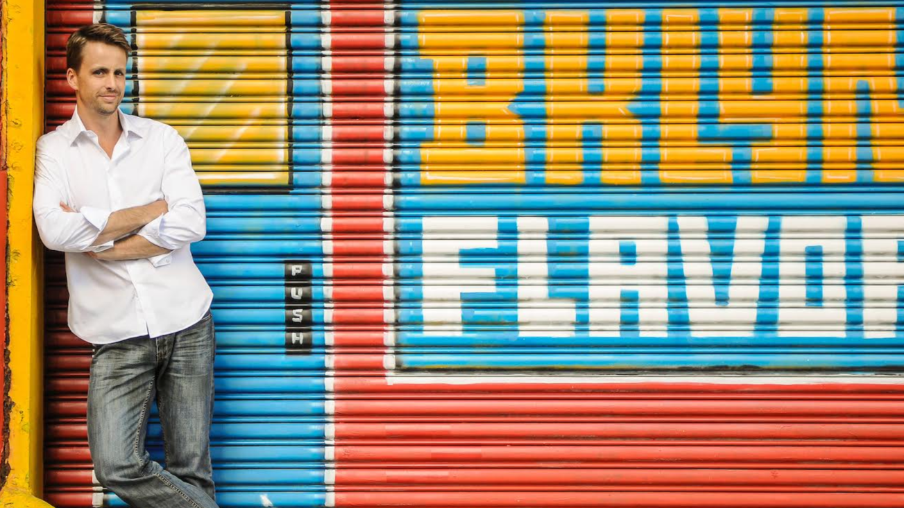 A man in a white shirt and gray jeans leaning against a colorful graffiti wall with a shutter painted in red, yellow, blue, black, and white, featuring the words 'TRUST' and 'SECURE'.