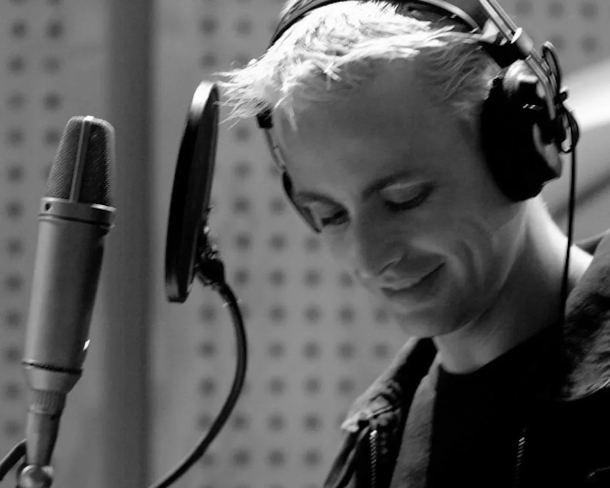 A young man with short light hair wearing headphones, standing in a recording studio, smiling as he looks down at a microphone in front of him.