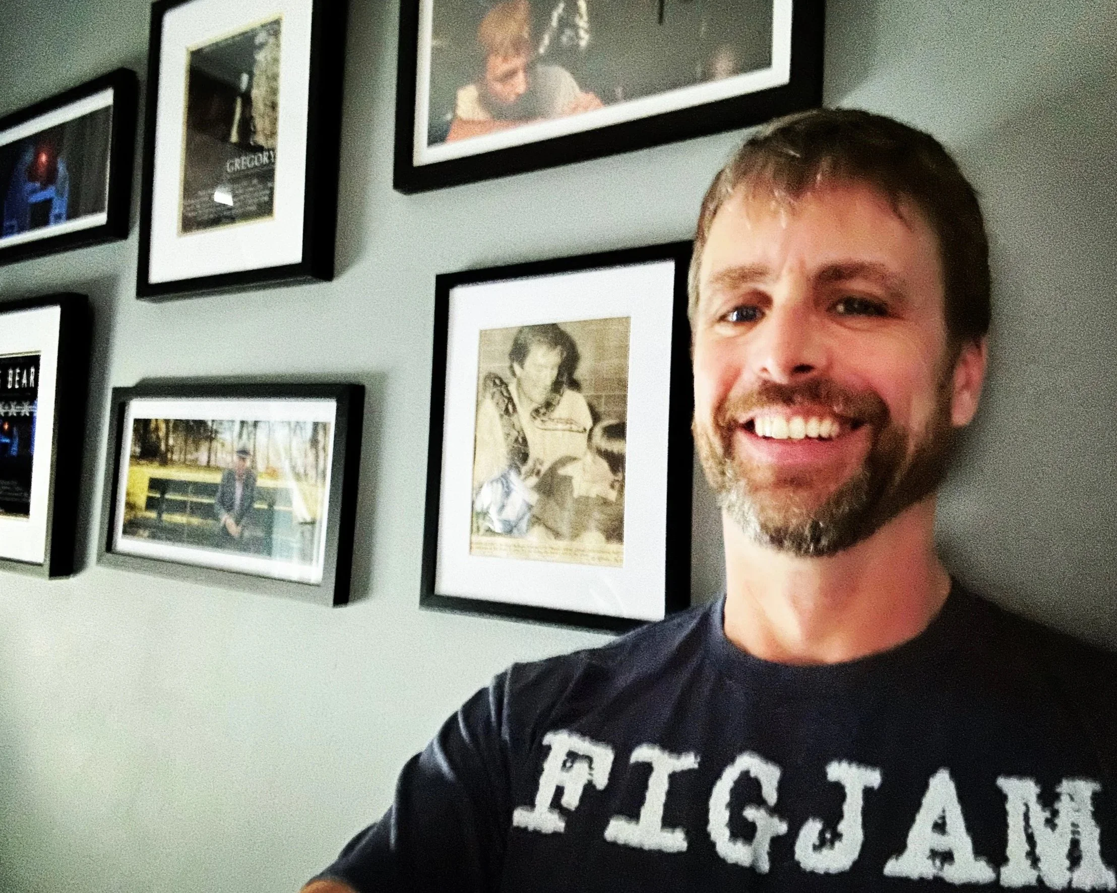 A smiling man with a beard taking a selfie in front of a wall decorated with framed photographs.