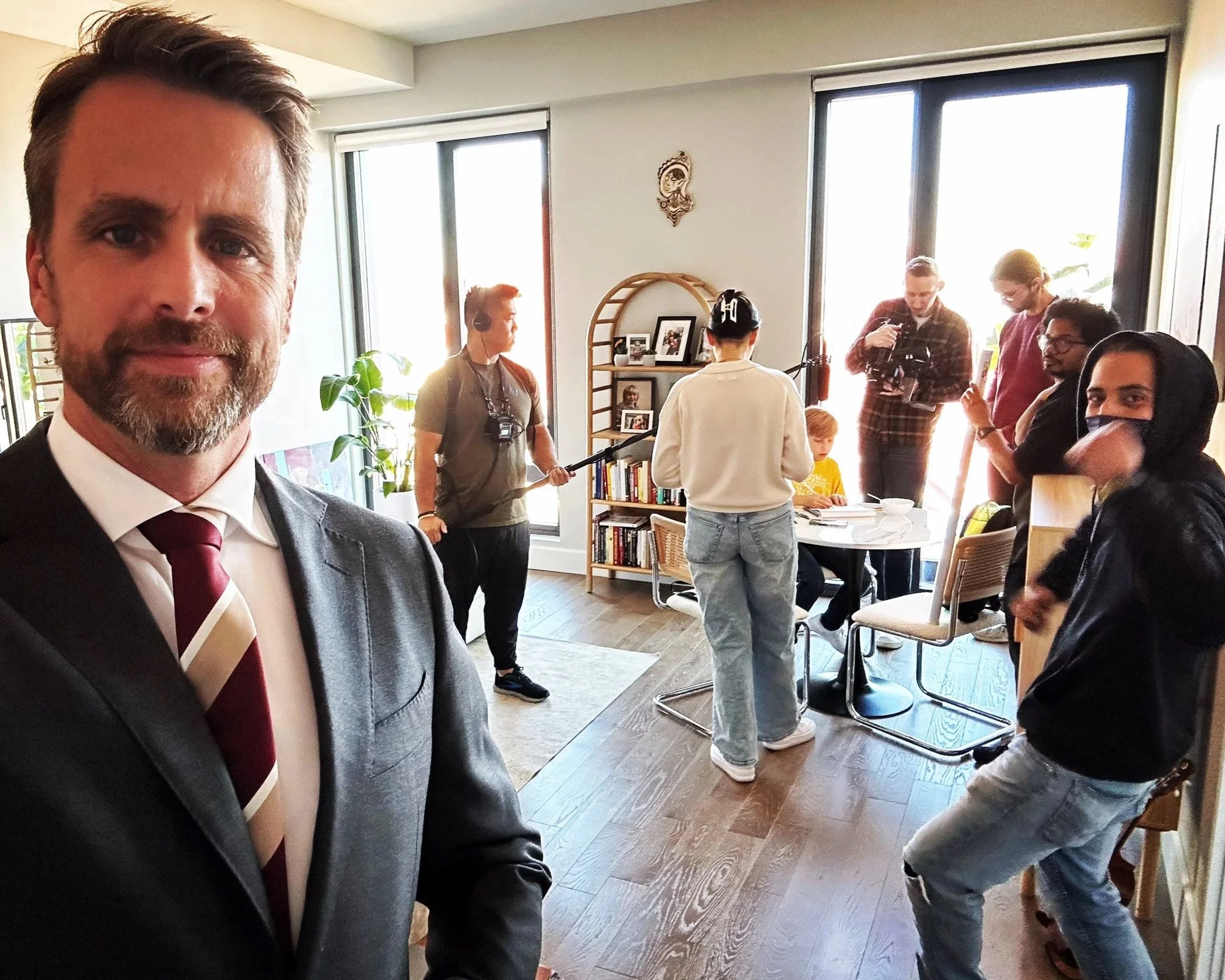 A man in a suit and tie taking a selfie in a well-lit room with a group of people engaging in filming and photography activities.