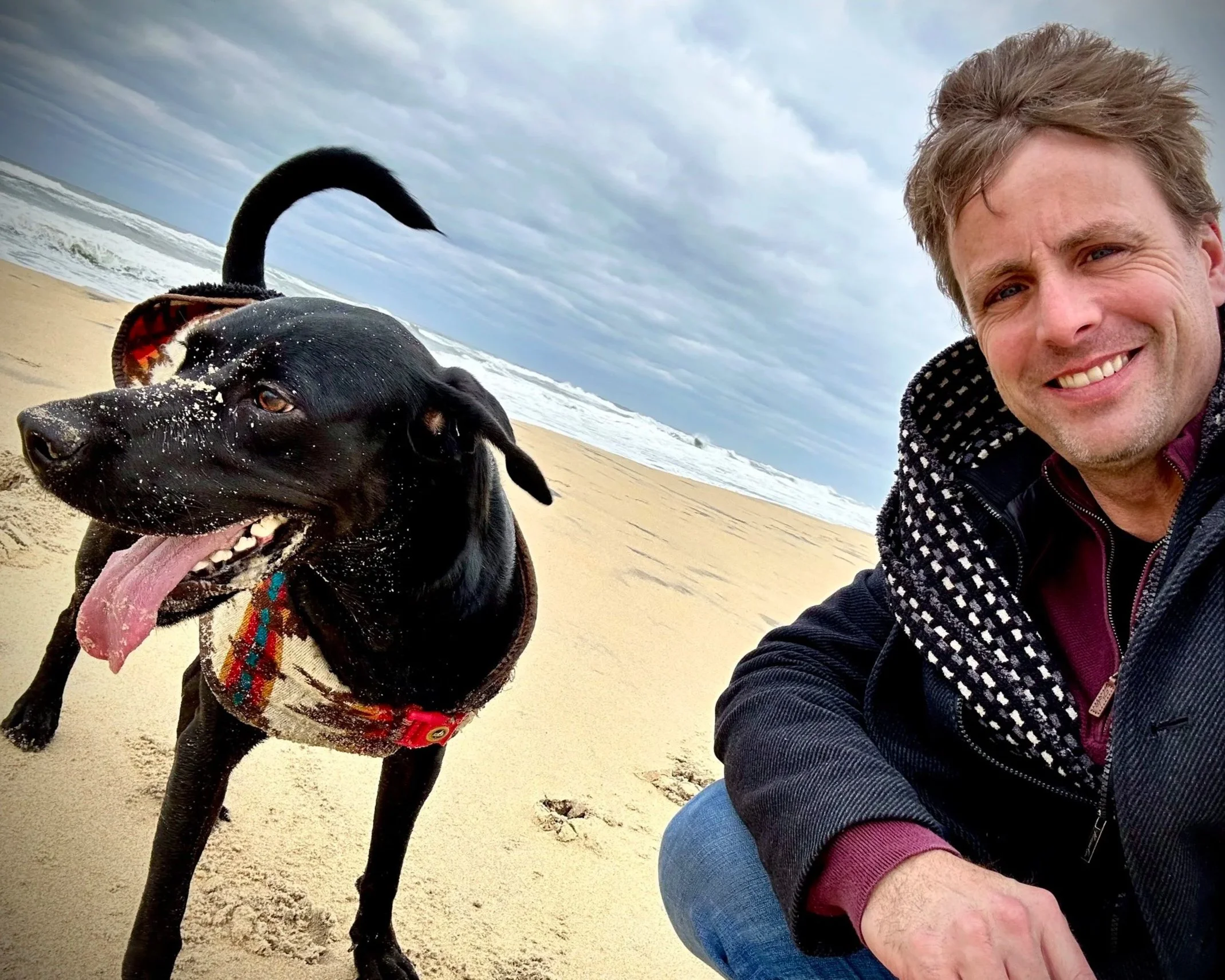 A man and a black dog taking a selfie on a beach with sand, ocean waves, and cloudy sky in the background.