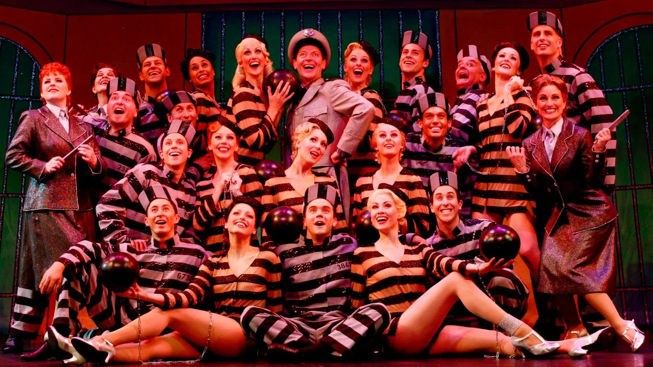 A theatrical cast in prison costumes on stage, with some holding balls and clubs, posing happily in front of metal prison bars set, with bright stage lighting.