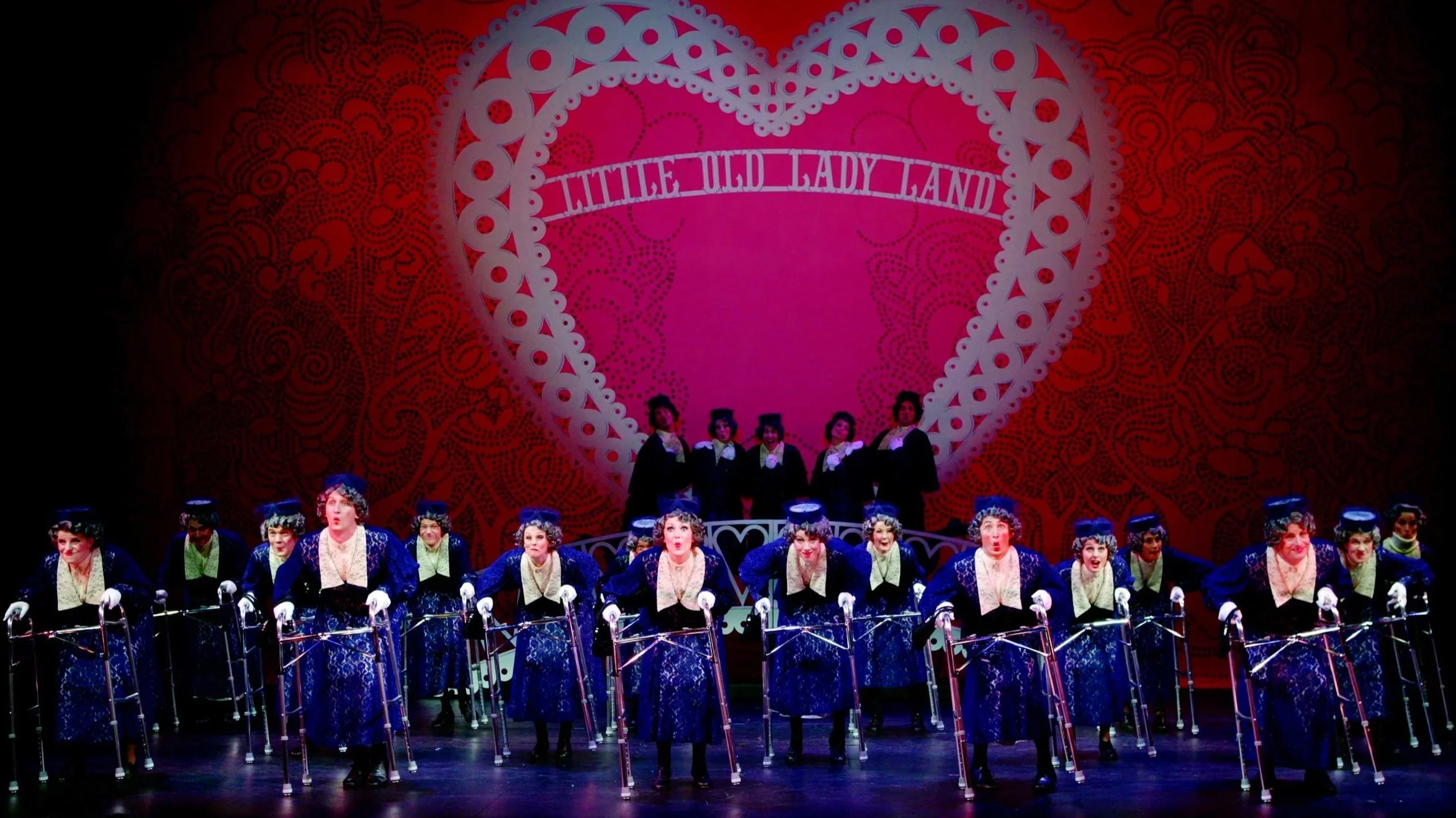 Stage performance of a chorus of women dressed in blue with crutches in front of a large pink heart backdrop with the words 'Little Old Lady Land'.