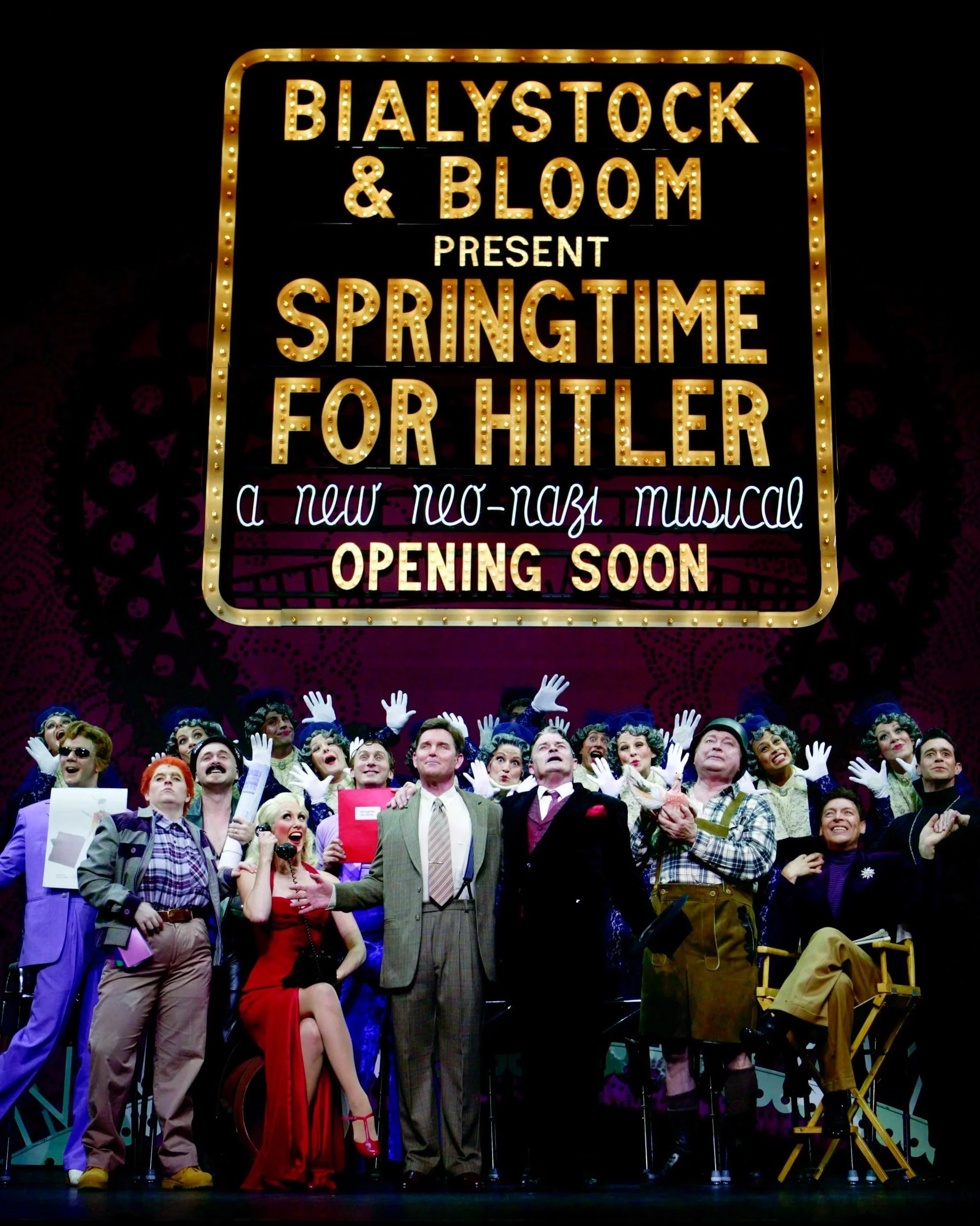 A group of actors on stage in costumes, participating in a theatrical performance, with a large, illuminated sign overhead promoting the show "Springtime for Hitler."