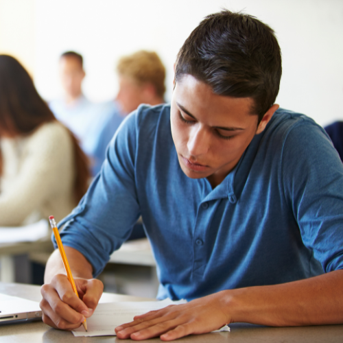 A young man in a classroom writing in a notebook with a yellow pencil.