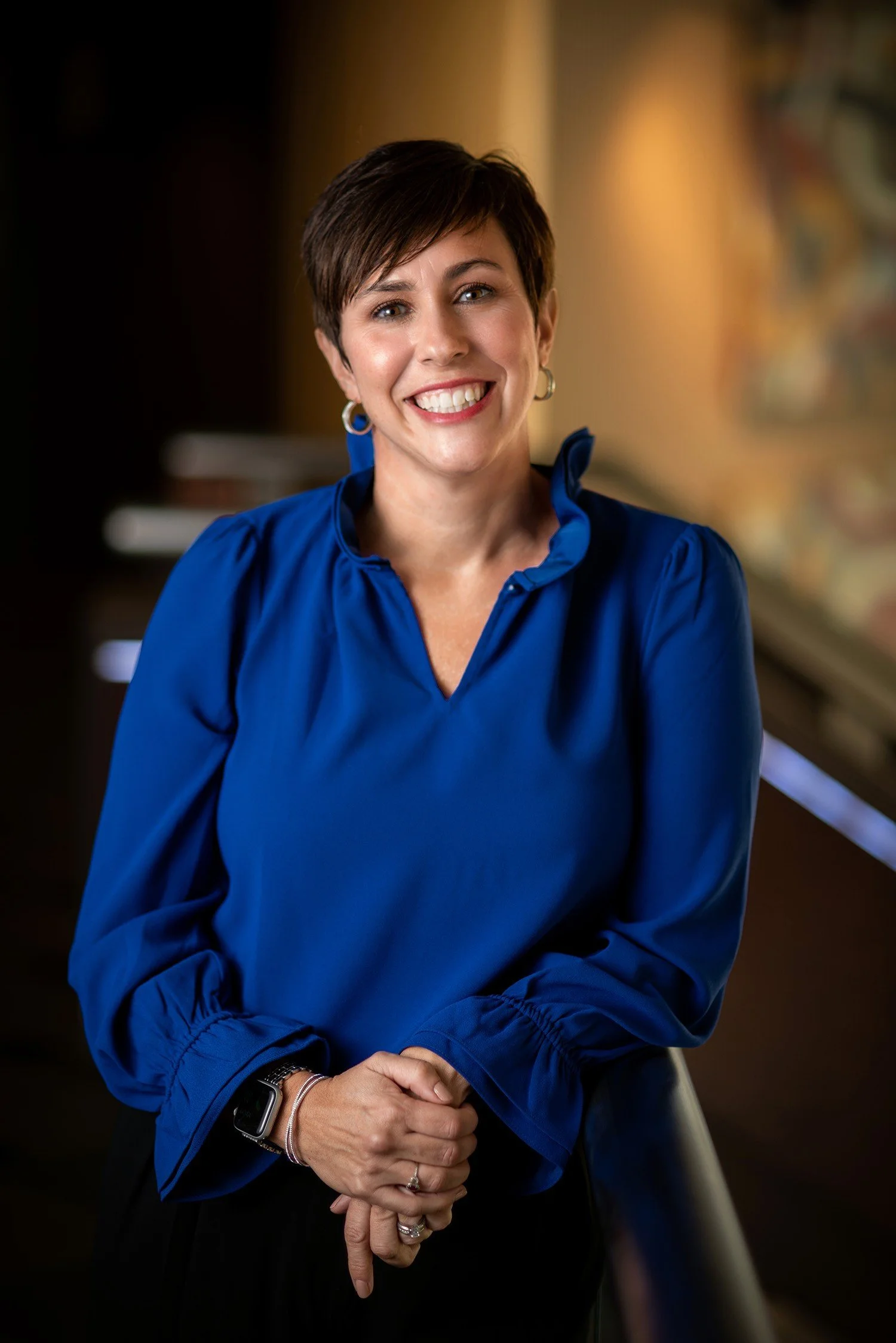 A woman with short brown hair wearing a bright blue blouse smiling and holding her hands together, standing indoors with a blurred background.