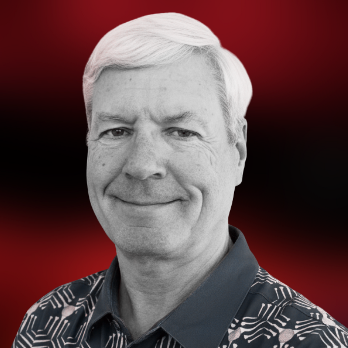 Black and white portrait of a smiling middle-aged man with gray hair, wearing a patterned shirt, against a red and black gradient background.