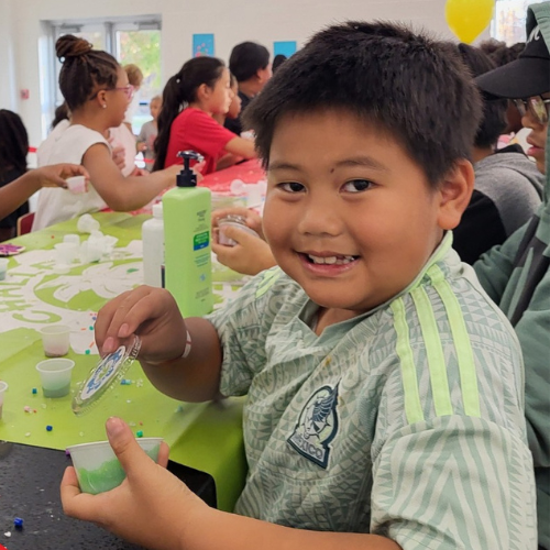 A young boy smiling at the camera while holding a small green container, seated at a table with others during a craft activity, with colorful decorations and supplies around.