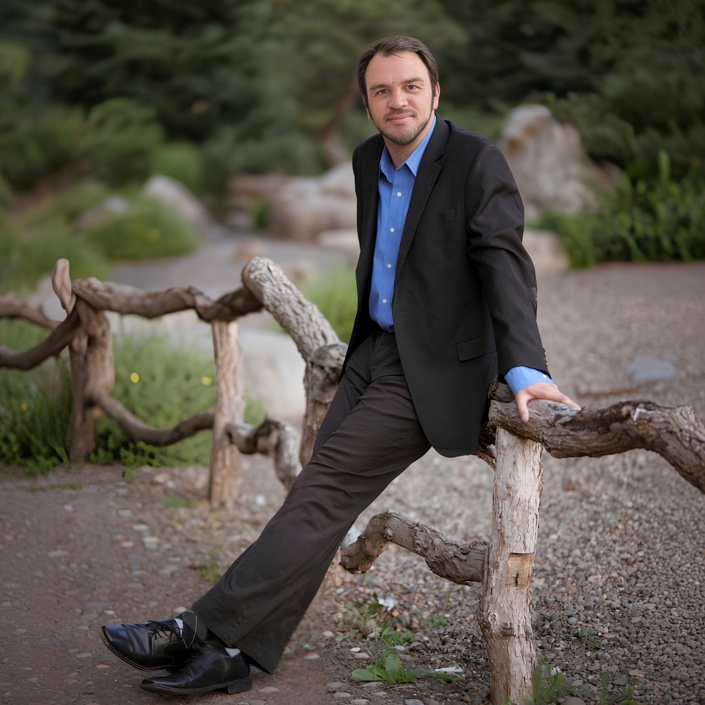 A man in a suit leaning against a rustic wooden fence in a green outdoor park setting.