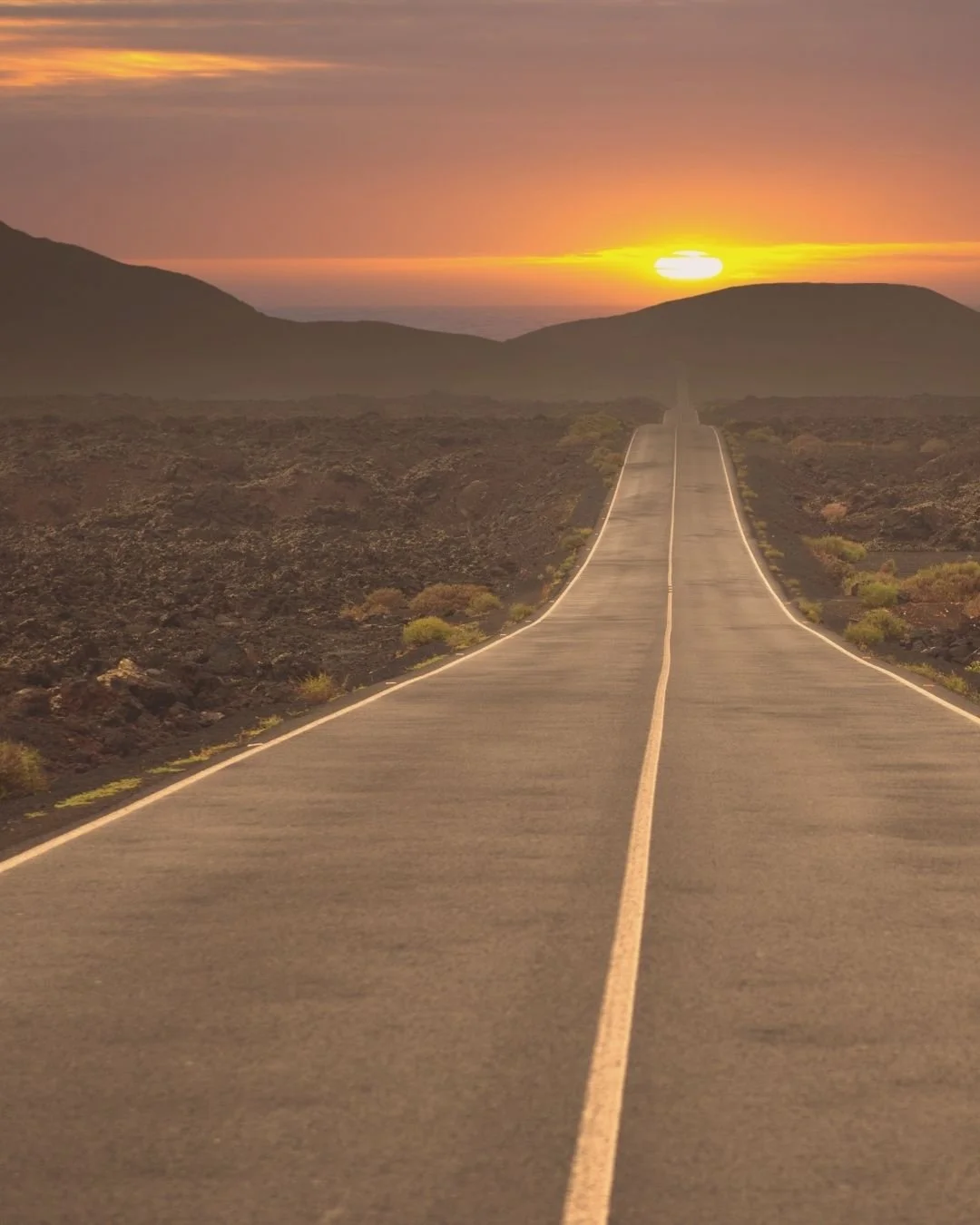 A long, straight road with yellow dividing lines extending into the horizon at sunset, surrounded by desert terrain with low shrubs and distant mountains under a colorful sky.