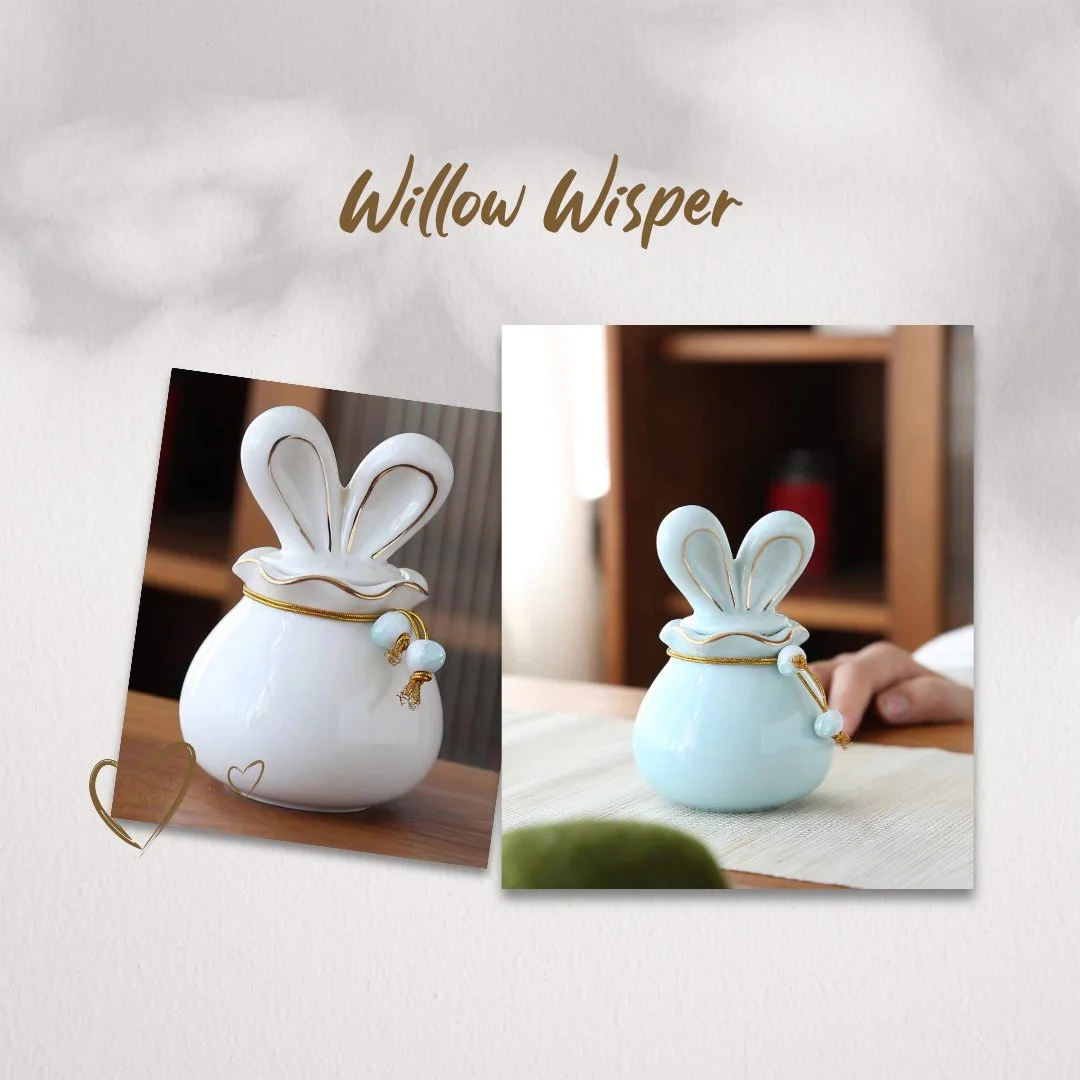 Two ceramic bunny-shaped vases with gold accents, labeled "Willow Whisper." One is white, placed on a wooden surface, and the other is light blue, set on a table with a blurred background.