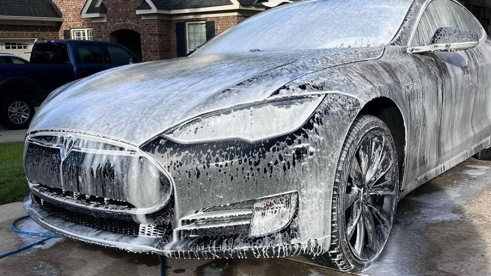 A silver Tesla car being washed with soap and water, covered in suds on the front and side, with a backyard and brick houses in the background.